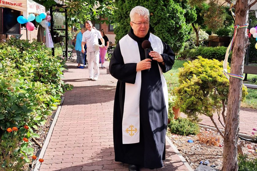 Modlitwa zakonnika – Zakonnik w czarno-białym habicie z mikrofonem w ręku prowadzi modlitwę podczas uroczystości plenerowej. W tle widać namiot i uczestników spotkania