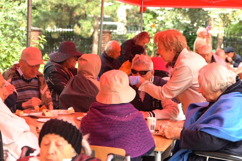 Seniorzy przy stole – Osoby starsze siedzące pod namiotem, ubrane w płaszcze i kapelusze, spożywają posiłek podczas pikniku