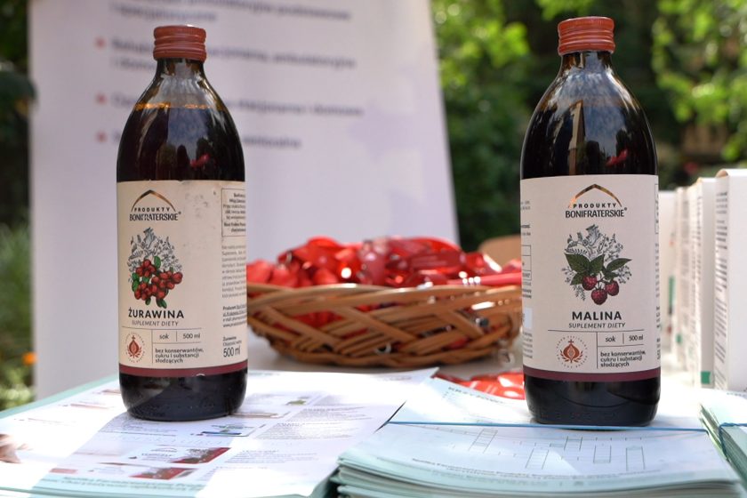 Produkty Bonifraterskie – soki – Zbliżenie na butelki soków żurawinowego i malinowego marki „Produkty Bonifraterskie”, ustawione na stole z materiałami informacyjnymi
