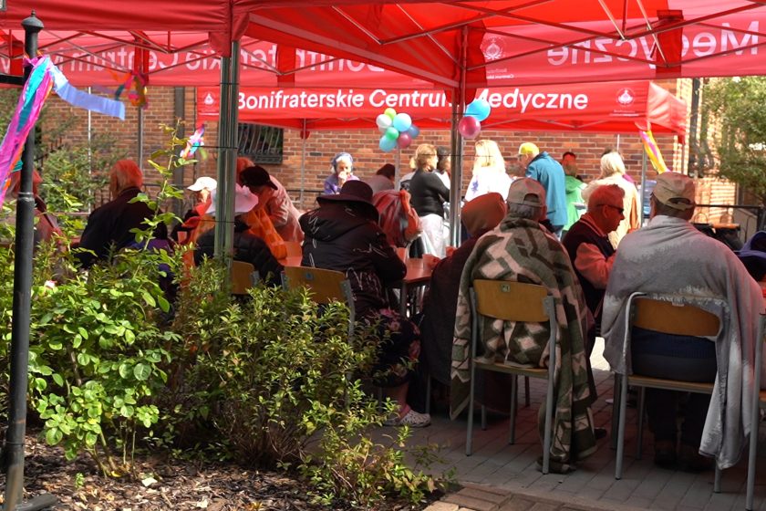 Piknik pod namiotami – Grupa osób siedzących przy stołach pod czerwonymi namiotami Bonifraterskiego Centrum Medycznego, w tle dekoracje i uczestnicy pikniku
