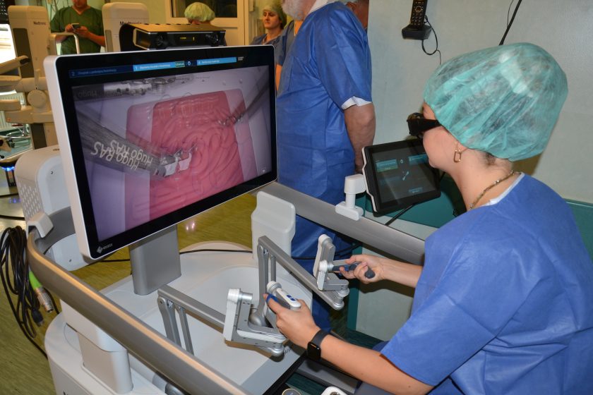 Osoba w niebieskim fartuchu chirurgicznym i czepku operacyjnym obsługuje konsolę sterującą robota Hugo RAS, patrząc na ekran z obrazem 3D wnętrza jamy brzusznej podczas symulacji zabiegu