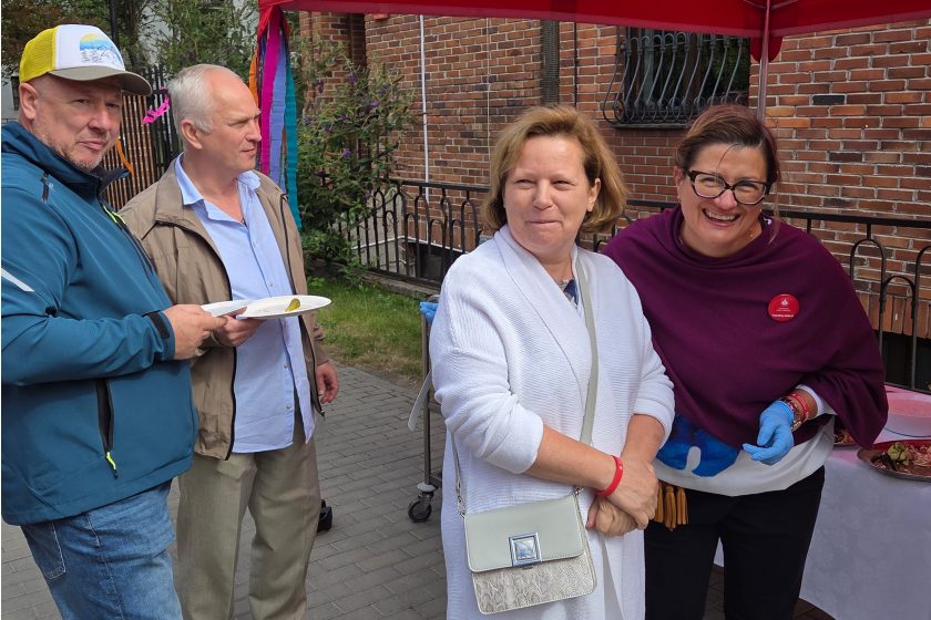 Rozmowy przy poczęstunku – Grupa uczestników wydarzenia przy stole z jedzeniem, dwie kobiety uśmiechają się do obiektywu, obok nich stoją mężczyźni z talerzami