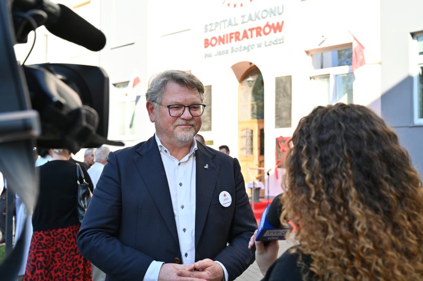 Mężczyzna w garniturze udziela wywiadu przed wejściem do Szpitala Zakonu Bonifratrów św. Jana Bożego w Łodzi, obok widoczny napis z nazwą placówki.