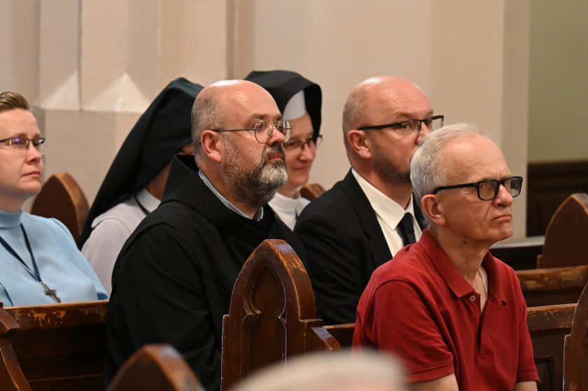 Zakonnik w habicie oraz wierni świeccy siedzą w ławkach kościelnych, wsłuchując się w przebieg nabożeństwa jubileuszowego