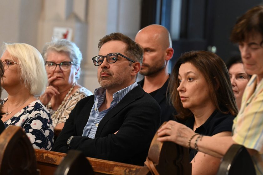 Grupa wiernych siedzących w ławkach w kościele podczas uroczystości. Na pierwszym planie mężczyzna w okularach i granatowej marynarce
