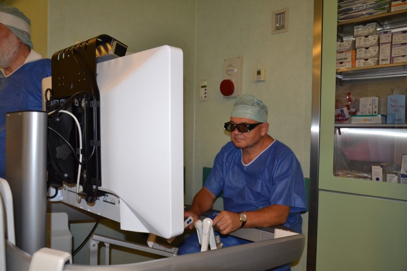 Lekarz w okularach 3D siedzi przy konsoli sterującej robotem chirurgicznym, koncentrując się na obrazie zabiegu wyświetlanym na dużym monitorze