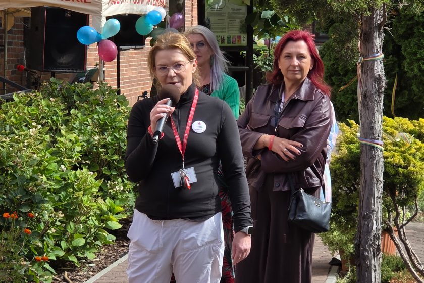 Przemówienie pracowniczki – Kobieta z mikrofonem i identyfikatorem Bonifraterskiego Centrum Medycznego przemawia podczas wydarzenia w ogrodzie. Za nią stoją uczestnicy spotkania