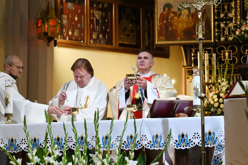 Kapłan w złotych szatach podnosi kielich podczas Eucharystii, obok niego dwaj księża w białych ornatach