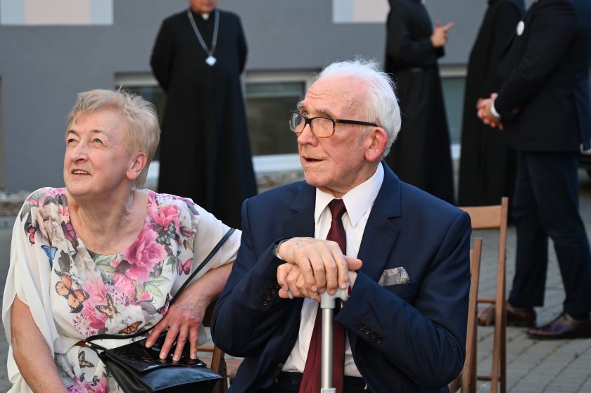 Starsze małżeństwo siedzi obok siebie podczas uroczystości jubileuszowych. Mężczyzna trzyma laskę, a kobieta ma na sobie sukienkę w kwiaty i motyle
