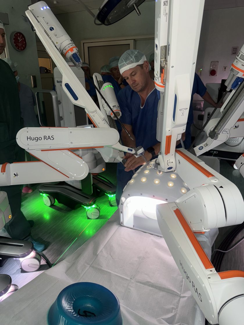 Lekarz w niebieskim fartuchu i czepku operacyjnym wykonuje symulację zabiegu, obsługując ramię robota Hugo RAS przy oświetleniu sali operacyjnej z zielonymi i białymi światłami