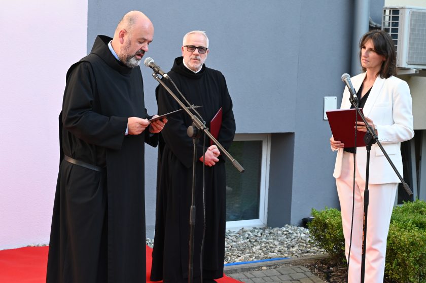 Zakonnik przemawia z kartki przy mikrofonie podczas uroczystości jubileuszowej, obok stoją inny zakonnik i kobieta w białym garniturze