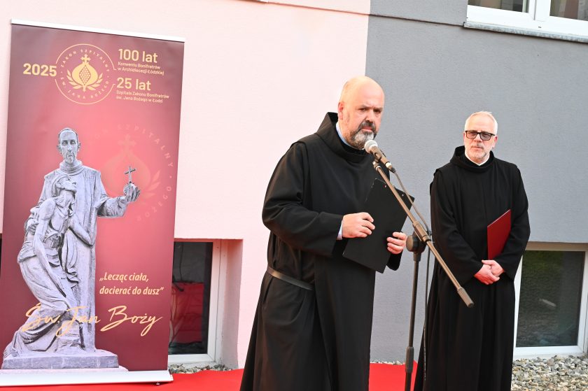 Zakonnik w czarnym habicie wygłasza przemówienie przy mikrofonie, obok baner jubileuszowy z wizerunkiem św. Jana Bożego