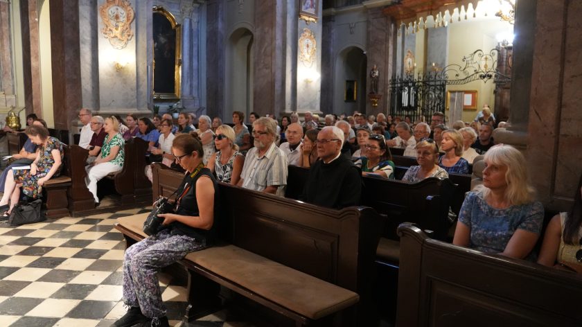 Pełny kościół; wierni siedzą w ławkach i słuchają koncertu. Opis rozszerzony: Ujęcie na lewe rzędy ławek – kilkadziesiąt osób w różnym wieku, część w okularach przeciwsłonecznych, niektórzy notują. Z przodu kobieta z aparatem na pasku trzyma lustrzankę na kolanach. W tle marmoryzowane filary, kute kraty kaplicy i złocone kartusze. Posadzka w biało-czarną szachownicę, światło ciepłe, koncert trwa