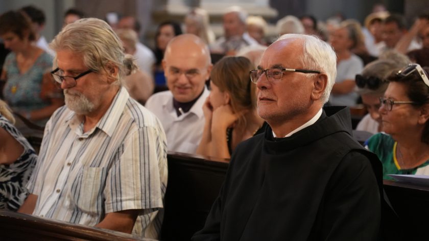 Zbliżenie na słuchaczy: zakonnik w czarnym habicie i mężczyzna w koszuli w paski. Opis rozszerzony: Pierwszy plan wypełniają twarze skupionych słuchaczy; po prawej siedzi starszy mężczyzna w habicie z kołnierzem, obok niego pan w jasnej, prążkowanej koszuli, z okularami zsuniętymi na czubek nosa. Głębia ostrości mała – tło zmiękczone, ale widać kolejne rzędy ław i rozświetlone profile słuchaczy