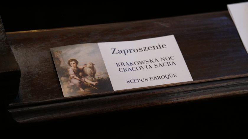 Zaproszenie „Krakowska Noc Cracovia Sacra – SCEPUS BAROQUE” na ławce kościelnej. Opis rozszerzony: Na ciemnym, politurowanym drewnie ławki leży biały kartonik z reprodukcją klasycznego obrazu (dziecko z barankiem). Po prawej druk: „Zaproszenie / KRAKOWSKA NOC CRACOVIA SACRA / SCEPUS BAROQUE”. Miękkie światło boczne podkreśla fakturę drewna i krawędzie kartonika