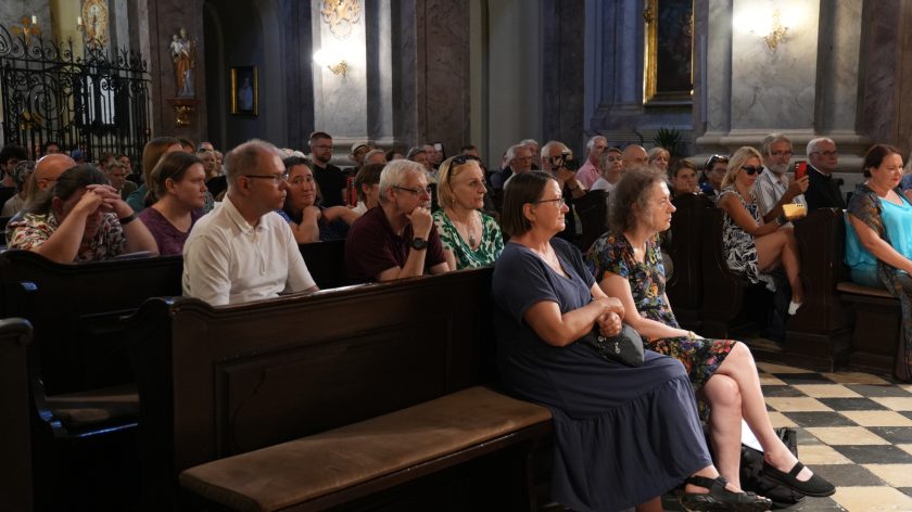 Słuchacze w półzbliżeniu; twarze skupione, w tle krata kaplicy i rzeźby. Opis rozszerzony: Kadr obejmuje lewy sektor ławek. Kilkanaście osób zwróconych ku scenie – niektórzy z rękami splecionymi, inni nagrywają telefonem. Światło z okien i lamp tworzy ciepłe refleksy na włosach i drewnie ławek. W głębi kuta krata i figura świętego na konsoli