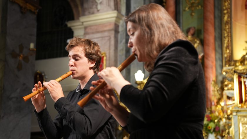 Muzycy na fletach prostych w trakcie koncertu, ujęcie z boku. Opis rozszerzony: Kadr uchwycił muzyków z perspektywy bocznej – oboje wpatrzeni w instrumenty, grają synchronicznie. Na dalszym planie widoczny fragment ściany kościoła z marmurowymi pilastrami i złotymi ozdobami. Gra świateł nadaje scenie uroczystego charakteru, a perspektywa bliskiego planu podkreśla ich zaangażowanie