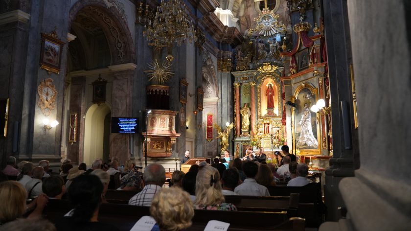 Szeroki kadr – koncert zespołu barokowego w kościele, widownia na ławkach. Opis rozszerzony: Zdjęcie obejmuje całą nawę kościoła z widokiem na prezbiterium. Przed ołtarzem głównym śpiewaczka i muzycy w czerni wykonują koncert, obok nich pozytyw. Ołtarz bogato zdobiony złoceniami, z figurami i obrazem Matki Bożej w srebrnej sukience. Publiczność wypełnia ławki, a nad sceną wisi ogromny żyrandol. Po lewej ekran z tekstami pieśni