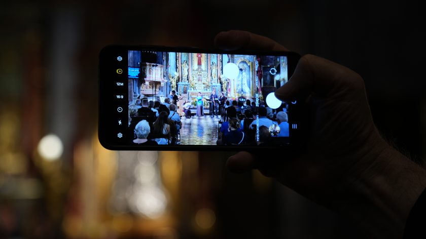 Widok koncertu przez ekran telefonu trzymanego w dłoni. Opis rozszerzony: Pierwszy plan zajmuje dłoń trzymająca smartfon, na którego ekranie widać dokładne ujęcie sceny koncertu: śpiewaczka, muzycy i ołtarz w tle. Sama sala i widownia pozostają lekko rozmyte, dzięki czemu ekran telefonu staje się centralnym punktem kadru. To ujęcie ukazuje nowoczesną perspektywę dokumentowania wydarzenia