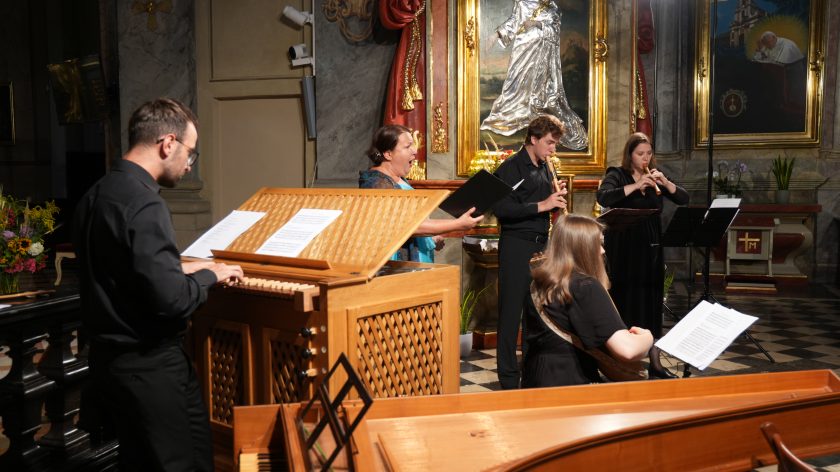 Zespół barokowy z wokalistką i organistą podczas występu w kościele. Opis rozszerzony: Na scenie pięcioro artystów – z lewej organista przy pozytywie, dalej śpiewaczka, obok niej dwóch flecistów i lutnistka. Wszyscy skupieni na muzyce, pulpity z nutami ustawione przed nimi. W tle monumentalny obraz Matki Bożej w srebrzystej sukience i złocone ramy ołtarza. Na pierwszym planie widoczny fragment klawesynu, co podkreśla bogactwo instrumentalne koncertu