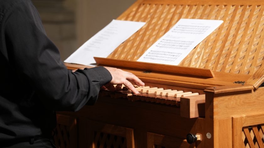 Zbliżenie na dłonie organisty grającego na pozytywie. Opis rozszerzony: Fotograf uchwycił dłonie muzyka naciskające drewniane klawisze małego pozytywu. Na pulpicie leżą zapisane nuty, a całość instrumentu ozdobiona jest geometryczną kratką z drewna. Światło punktowe wydobywa fakturę klawiatury i delikatny ruch palców. To ujęcie intymnie oddaje moment pracy instrumentalisty