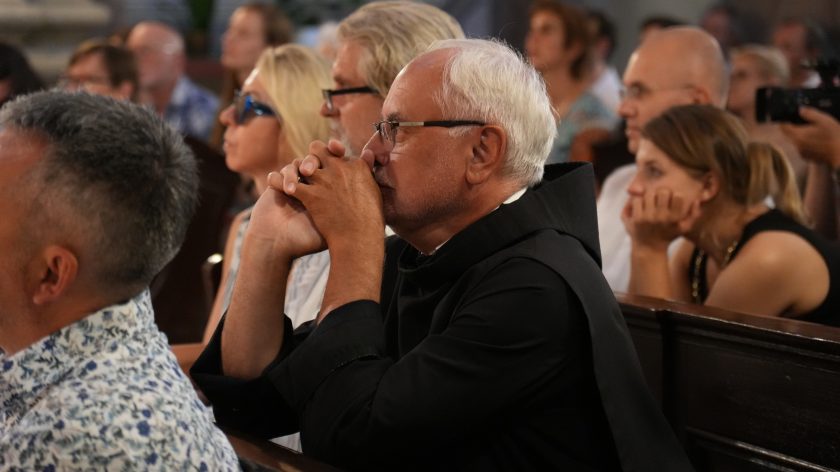 Zakonnik w habicie siedzi w ławce kościelnej, pogrążony w skupieniu i modlitewnej zadumie. Jego dłonie złożone są przy ustach, a spojrzenie skierowane ku ołtarzowi. W tle widoczni wierni, którzy w ciszy wsłuchują się w muzykę