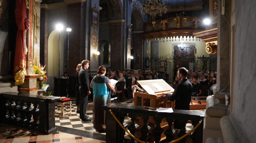 Ujęcie sceniczne – muzycy zespołu wykonują utwór podczas koncertu. Organista gra na pozytywie, a pozostali artyści stoją w pobliżu, śpiewając i grając dla zgromadzonej publiczności. W tle widać wnętrze świątyni wypełnione słuchaczami