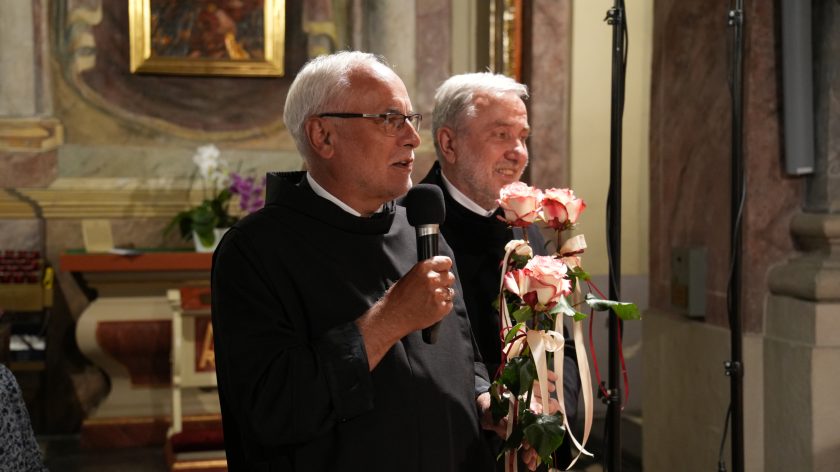 Brat zakonny w habicie dziękuje artystom, trzymając w dłoni mikrofon oraz bukiet róż. Obok niego stoi drugi brat, uśmiechnięty, w tle widoczna złocona dekoracja ołtarza bocznego