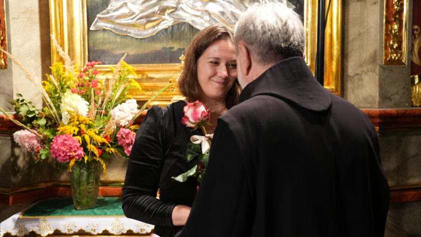 Ujęcie indywidualne – zakonnik wręcza różę jednej z artystek. Kobieta w czarnej sukni uśmiecha się wdzięcznie, przyjmując kwiat na tle bogato zdobionego ołtarza bocznego z wazonem pełnym kwiatów