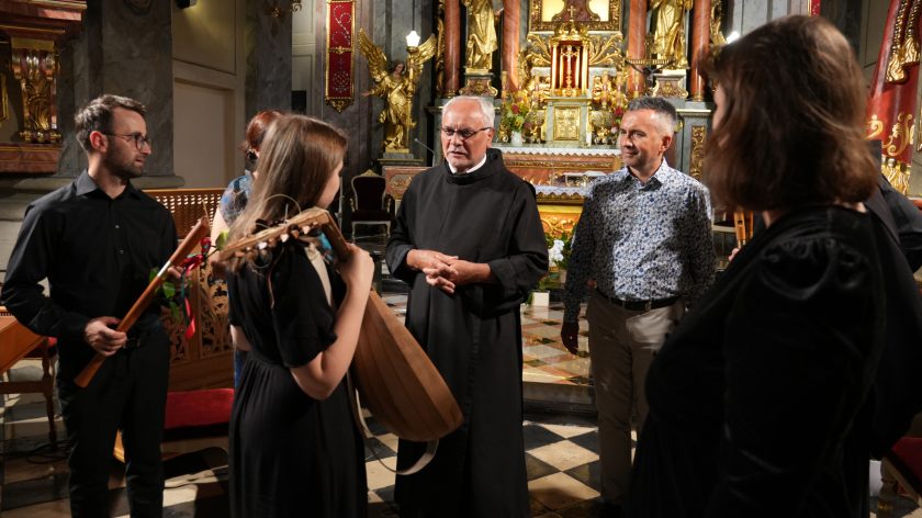 Rozmowa po koncercie – brat zakonny w habicie rozmawia z lutnistką, która trzyma swój instrument. Wokół stoją inni muzycy i uczestnicy spotkania, a w tle widoczny jest ołtarz oraz elementy barokowego wystroju kościoła