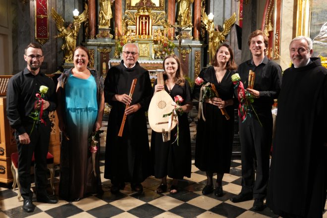 Grupa artystów zespołu Scepus Baroque wraz z braćmi bonifratrami po koncercie w prezbiterium kościoła. Muzycy trzymają w dłoniach instrumenty – lutnię i flety – oraz otrzymane kwiaty. W tle widoczny bogato zdobiony ołtarz główny z figurami aniołów i złoceniami