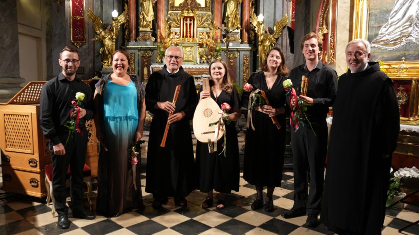 Grupa artystów zespołu Scepus Baroque wraz z braćmi bonifratrami po koncercie w prezbiterium kościoła. Muzycy trzymają w dłoniach instrumenty – lutnię i flety – oraz otrzymane kwiaty. W tle widoczny bogato zdobiony ołtarz główny z figurami aniołów i złoceniami