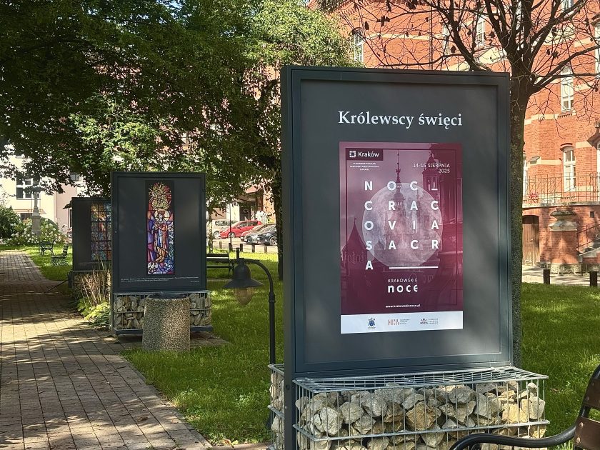 Plansza wystawowa z napisem „Królewscy święci” i plakatem wydarzenia Noc Cracovia Sacra, 14–15 sierpnia 2025, a w tle kolejne fotografie witraży umieszczone wzdłuż alejki ogrodowej