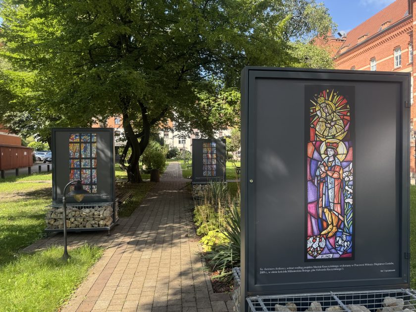 Alejka w cieniu drzew prowadząca przez teren zielony, po obu stronach stoją plansze z reprodukcjami barwnych witraży. Na pierwszym planie przedstawienie św. Kazimierza Królewicza w koronie, z aureolą i modlitewnie złożonymi rękami
