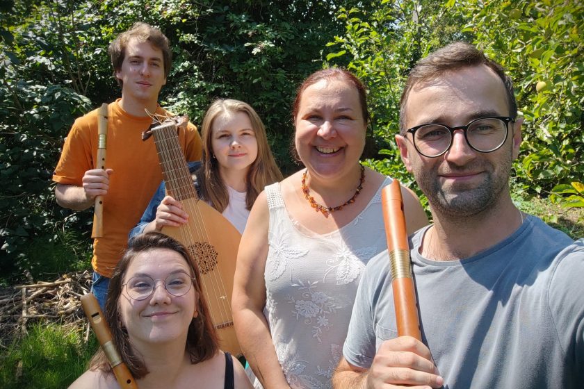 Zdjęcie grupowe zespołu Scepus Baroque Pięć osób stoi na tle zieleni w słoneczny dzień. Na pierwszym planie od lewej: kobieta w okularach trzymająca flet, mężczyzna w okularach trzymający flet prosty. W drugim rzędzie: młody mężczyzna w pomarańczowej koszulce z fletem, młoda kobieta z lutnią oraz kobieta w białej bluzce, uśmiechnięta, w naszyjniku z bursztynu. Wszyscy patrzą w stronę obiektywu