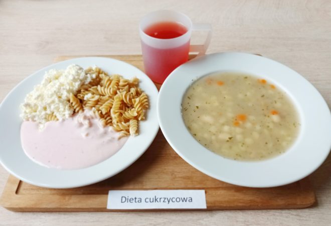Zdjęcie przedstawia zestaw obiadowy dla diety cukrzycowej. Na białym talerzu po lewej stronie znajdują się gotowany makaron świderki, biały twaróg i sos truskawkowy lub jogurtowy. Na drugim talerzu: zupa jarzynowa z ziemniakami, marchewką i przyprawami ziołowymi. W tle przezroczysty kubek z czerwonym kompotem. Całość ustawiona na drewnianej tacy z etykietą „Dieta cukrzycowa”