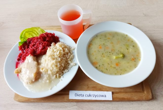 Na drewnianej tacy ustawiono obiad: na jednym talerzu porcja gotowanego kurczaka w sosie, kasza jęczmienna, buraczki tarte oraz liść sałaty. Obok, w głębokim talerzu, znajduje się zupa jarzynowa z brokułami, marchewką i ziemniakami. Do picia w przezroczystym kubku z uchem podano czerwony napój. Na tacy widoczny jest napis „Dieta cukrzycowa”