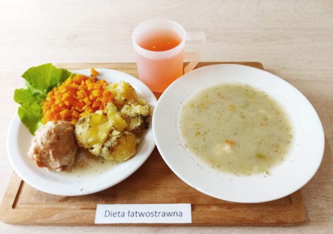 Obiad w diecie lekkostrawnej: gotowany pulpet z mięsa drobiowego, gotowane ziemniaki z koperkiem oraz marchewka pokrojona w kostkę, ułożone na liściu sałaty. Obok talerza miska z delikatną zupą jarzynową oraz różowy napój w plastikowym kubku. Całość podana na drewnianej tacy z kartką: „Dieta łatwostrawna”
