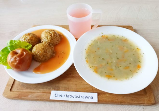 Zestaw obiadowy w diecie łatwostrawnej podany na białym talerzu: trzy kulki z kaszy jęczmiennej lub ryżowej w lekkim sosie pomidorowym, upieczone jabłko oraz liść sałaty. Obok znajduje się talerz z zupą jarzynową o kremowej konsystencji, z widocznymi kawałkami marchwi i ziół. Całość uzupełnia różowy napój w przezroczystym kubku. Zestaw ustawiony na drewnianej tacy z podpisem: „Dieta łatwostrawna”