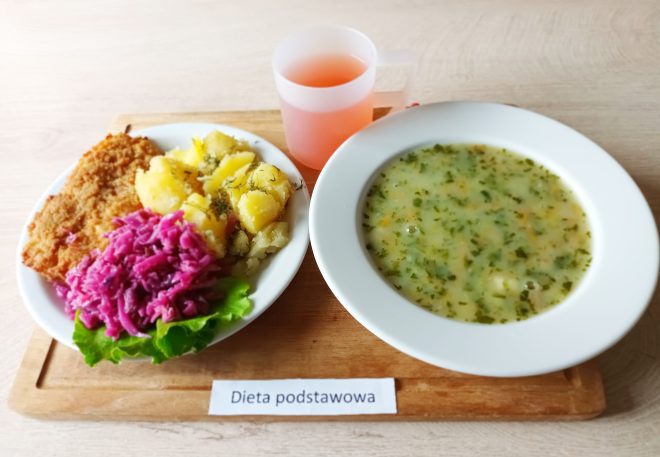 Dieta podstawowa: Drewniana taca z obiadem – na talerzu panierowany kotlet, gotowane ziemniaki z koperkiem oraz surówka z czerwonej kapusty na liściu sałaty; obok talerz z zupą jarzynową z ziemniakami i natką pietruszki oraz kubek z różowym kompotem. Na tacy karteczka z napisem „Dieta podstawowa”