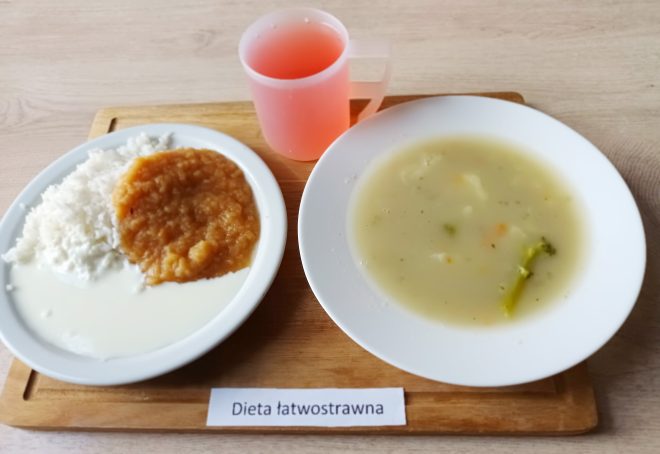 Na tacy ustawione są dwa białe talerze: w jednym zupa jarzynowa o kremowej konsystencji z widocznymi kawałkami brokuła i marchewki, w drugim ryż polany sosem mlecznym oraz porcja musu jabłkowego. Obok znajduje się różowy napój w plastikowym kubku