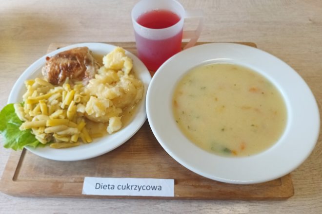 Dieta cukrzycowa. Na drewnianej tacy ustawiony zestaw obiadowy składający się z talerza z porcją ziemniaków z sosem, gotowaną żółtą fasolką szparagową i kawałkiem pieczonego mięsa drobiowego. Obok miska z zupą jarzynową o jasnym, kremowym kolorze oraz kubek z czerwonym kompotem