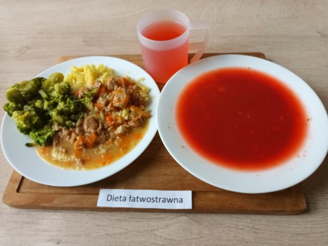 Na białym talerzu porcja gotowanych brokułów, makaronu oraz duszonego mięsa w sosie z warzywami. Obok znajduje się talerz z zupą pomidorową oraz przezroczysty kubek z czerwonym napojem. Pod talerzami leży kartka z napisem „Dieta łatwostrawna”