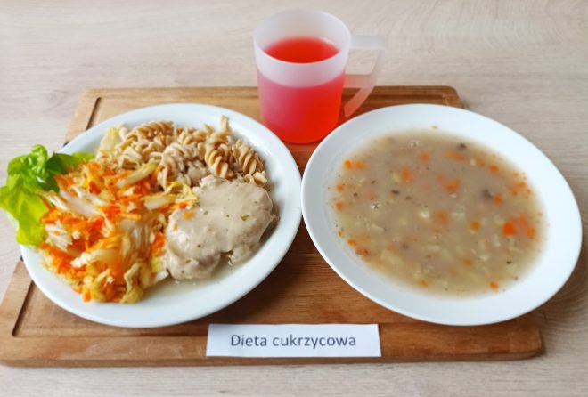 Na tacy podano dwudaniowy obiad w ramach „Diety cukrzycowej”: talerz z zupą jarzynową oraz drugie danie składające się z makaronu pełnoziarnistego, duszonego mięsa w sosie, surówki z kapusty i marchewki, a także kawałka sałaty. Do posiłku dodano kubek z czerwonym napojem owocowym