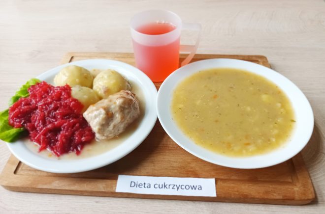 Na drewnianej tacy z karteczką „Dieta cukrzycowa” podano obiad. Po lewej stronie na białym talerzu znajduje się gotowany ziemniak, kawałek mięsa w jasnym sosie, surówka z buraków oraz liść sałaty. Obok stoi przezroczysty kubek z czerwonym napojem. Po prawej stronie znajduje się talerz z zupą jarzynową o kremowej konsystencji, z widocznymi kawałkami warzyw