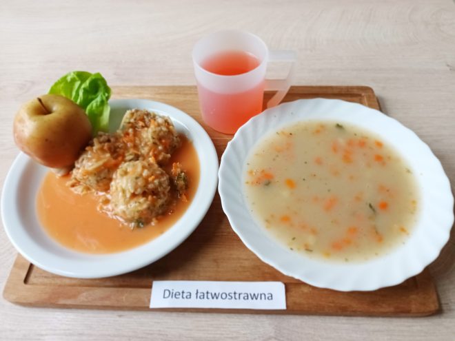 Na drewnianej tacy ustawiono posiłek w ramach diety łatwostrawnej. Widać głęboki talerz z zupą jarzynową o lekkiej, jasnej barwie z kawałkami marchewki i zieleniny. Obok, na mniejszym talerzu, znajdują się pulpety w sosie pomidorowym z dodatkiem listka sałaty i jabłko. Do zestawu dołączono przezroczysty kubek z różowym napojem owocowym