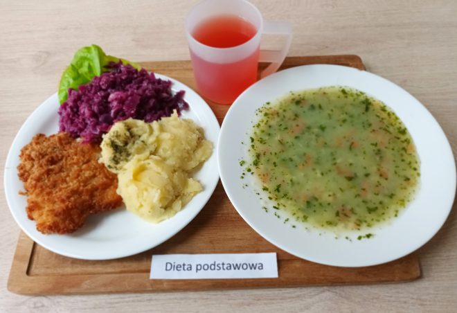 Na zdjęciu widoczny jest zestaw obiadowy w diecie podstawowej. Na talerzu znajdują się ziemniaki puree posypane koperkiem, panierowany kotlet schabowy oraz duszona czerwona kapusta z listkiem sałaty. Obok podano talerz z lekką zupą jarzynową z natką pietruszki. Do zestawu dołączono kubek z czerwonym kompotem owocowym. Posiłek ułożony jest na drewnianej tacce z podpisem „Dieta podstawowa”