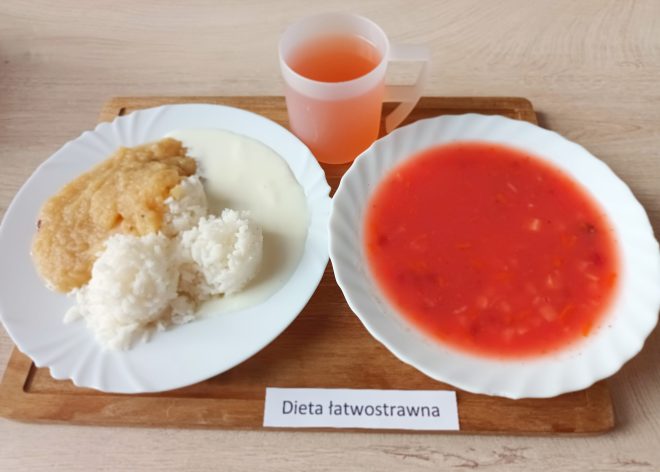 Na białym talerzu porcja białego ryżu z sosem i musem jabłkowym, obok talerz z zupą pomidorową. Do posiłku podano napój owocowy w plastikowym kubku. Całość ustawiona na drewnianej tacy z napisem „Dieta łatwostrawna”