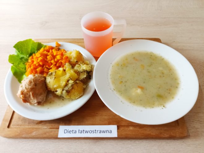 Talerz z porcją gotowanych ziemniaków posypanych koperkiem, gotowaną marchewką w kostkę, kotletem mielonym i liściem sałaty. Obok miska z zupą jarzynową na białym talerzu oraz kubek z czerwonym napojem. Na tacy napis: „Dieta łatwostrawna”
