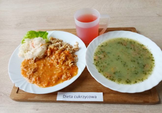 Na drewnianej tacy ułożono posiłek opisany jako „Dieta łatwostrawna”. Na talerzu znajduje się pieczywo: bułka pszenna i kromka chleba, a także plasterki wędliny drobiowej, ser twarogowy, kawałek masła, sałata oraz plasterki pomidora. Obok podano biały kremowy zupopodobny posiłek w głębokim talerzu oraz herbatę w przezroczystym kubku